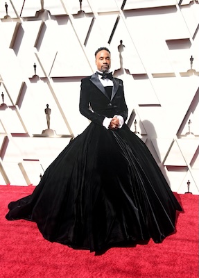 Billy Porter on a red carpet in front of a white wall, wearing a black gown that mimics a tuxedo on the top and widens out considerably on the bottom